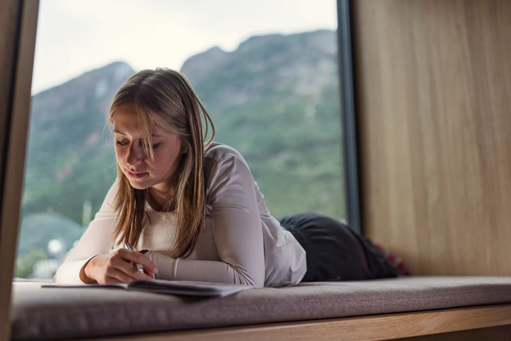 Woman writing in a notebook by a window with mountain view