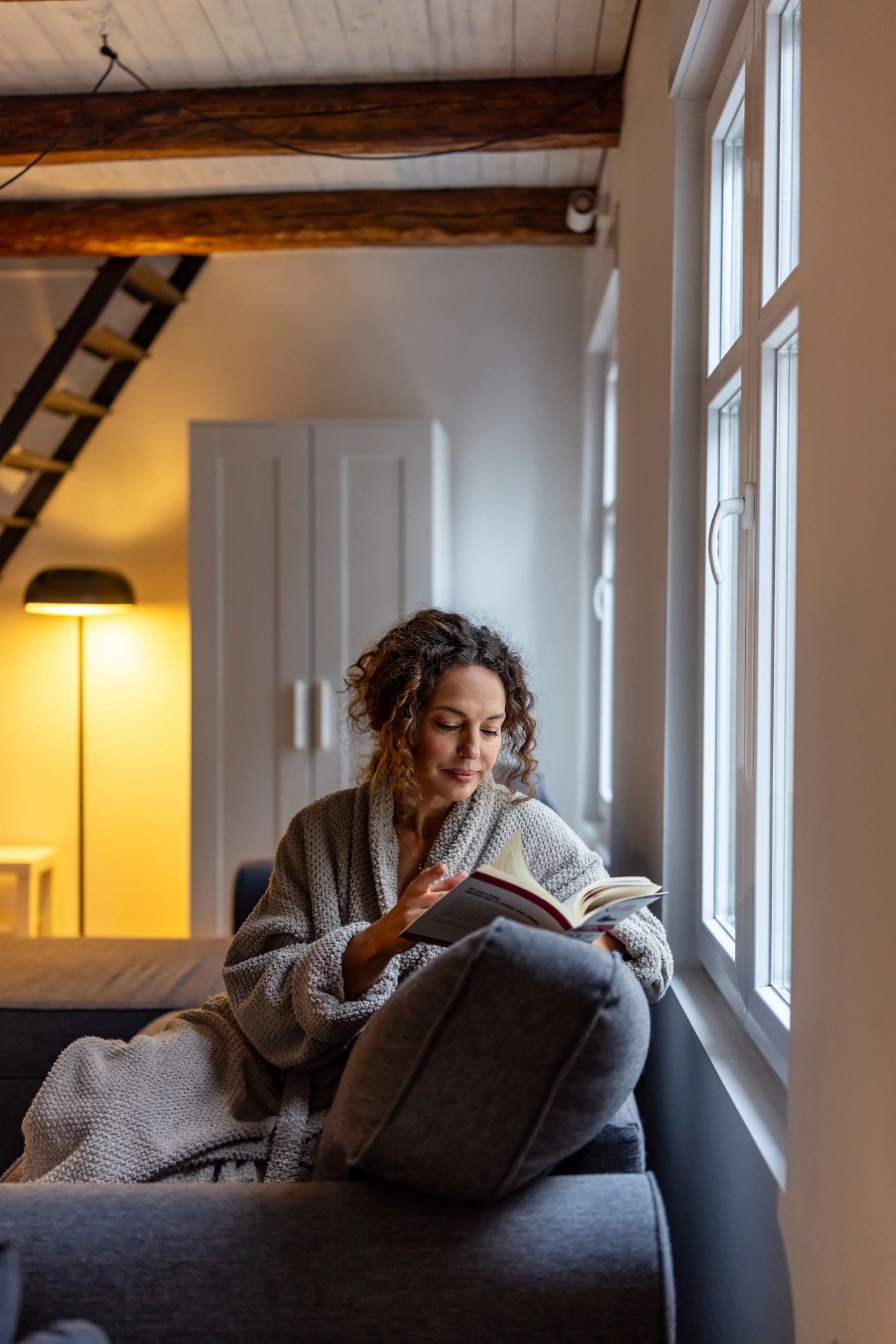 Woman relaxing by a bright window in a cozy living room, reading in soft natural light